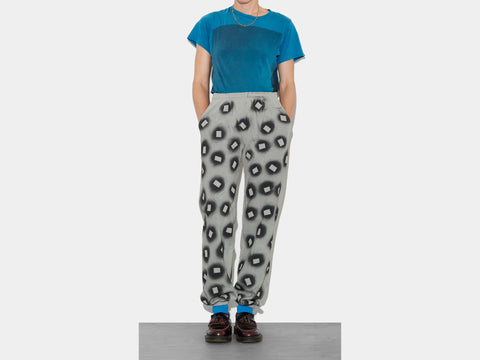 Female model wears 100% cotton sage sweatpants, perfectly cut, with hand-painted, black, negative cut-out squares extending across body and down legs. High-contrast composition really pops. The pattern continues on the other side of the pants. Pants are paired with blue t-shirt, blue socks, loafers and a gold necklace.