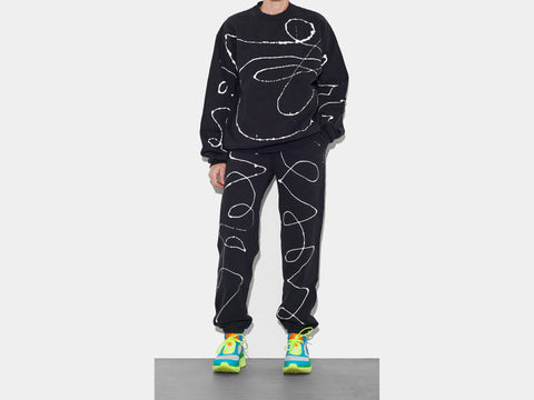 Model is shown wearing 100% cotton black sweatsuit, perfectly cut, which has scrawled, drippy white hand-painted line running across the body, sleeves, and down each leg.  